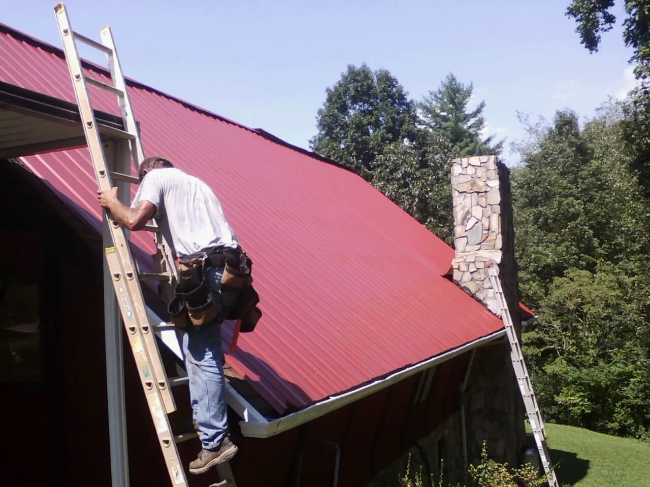 Roofer on a ladder beside a red metal roof and stone chimney — Soffit & Fascia Repair by Roofing Brothers in Myersville
